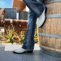 Person wearing blue jeans and white cowboy boots leaning against a wooden barrel.