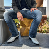 Person wearing blue jeans and white cowboy boots sitting on a wooden stool outdoors.